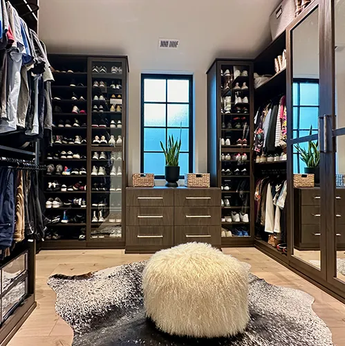 A luxurious walk-in closet by PNW Closets featuring dark wood custom cabinetry, integrated shoe racks with glass doors, a window seat with drawers, and a stylish shag rug with a plush ottoman.